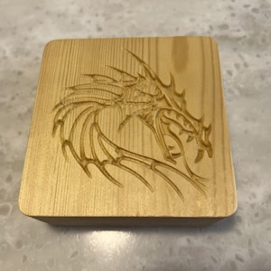 May include: A wooden square coaster with a carved dragon design. The dragon is facing right and has detailed wings and scales.