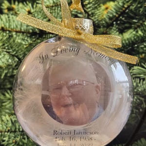 Può includere: Un addobbo natalizio in vetro trasparente con un nastro dorato e una foto di una persona all'interno. L'addobbo ha il testo "In Loving Memory" e "Robert Jamieson Feb. 16, 1958 - July 11, 2023" stampato su di esso.
