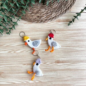 May include: Three handmade crochet seagull keyrings. Each seagull is white with grey wings, orange beaks and feet, and a different coloured hat: yellow, red, and purple. The keyrings are on a wooden surface with a woven basket and greenery in the background.
