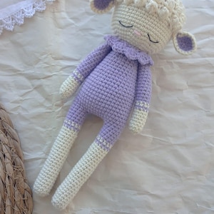 May include: A crocheted stuffed lamb toy with a white body and purple clothing. The lamb has a white face with closed eyes and a purple collar.