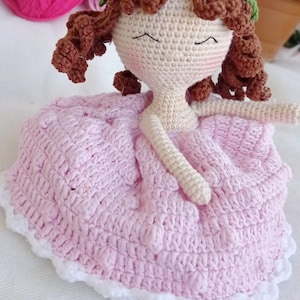 Puede incluir: Una muñeca de crochet con cabello castaño, un vestido rosa y una corona de flores. La muñeca tiene una expresión dulce y está sentada sobre una superficie blanca.