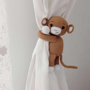 May include: A brown crocheted monkey toy with white accents is holding a white curtain with its arms. The monkey has a long tail and is holding the curtain closed.