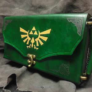 May include: A green leather clutch with a gold Triforce symbol and a brass clasp. The clutch has a black leather strap with a brass buckle.