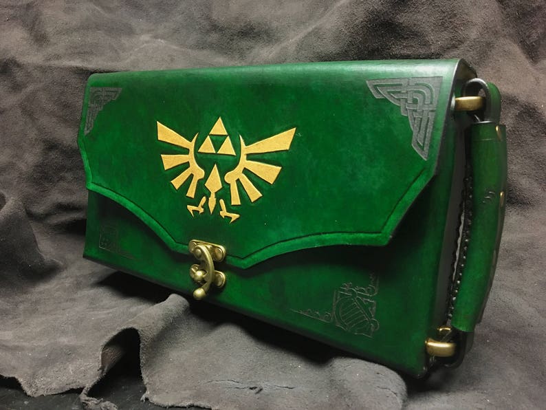 May include: A green leather clutch with a gold Triforce symbol and a brass clasp. The clutch has a black leather strap with a brass buckle.