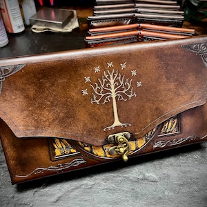 Op de afbeelding: Een bruin leren dagboek met een klep sluiting en een messing sluiting. Het dagboek heeft een reliëf ontwerp van een boom met een witte omtrek en sterren. Het dagboek ligt op een donker oppervlak.