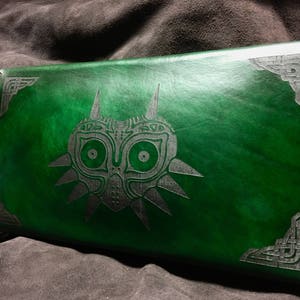 May include: A green leather notebook with a silver embossed design of the Majora's Mask from the Legend of Zelda video game series. The notebook has a black leather trim.