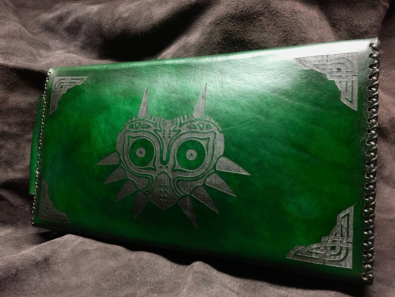 May include: A green leather notebook with a silver embossed design of the Majora's Mask from the Legend of Zelda video game series. The notebook has a black leather trim.