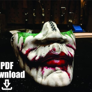 Pode incluir: Uma máscara de couro branco com tinta verde e vermelha, que se assemelha a um sorriso estilo Joker. A máscara tem rebites de metal e detalhes de costura.