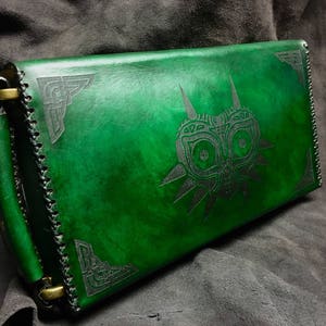 May include: A green leather clutch with a black embossed design of a mask with a triangular shape and a stylized face. The clutch has a flap closure and a brass buckle.
