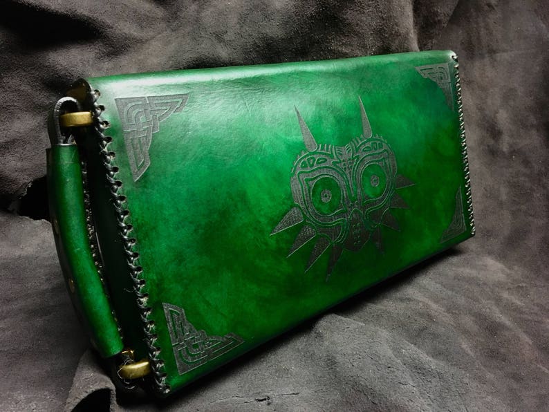 May include: A green leather clutch with a black embossed design of a mask with a triangular shape and a stylized face. The clutch has a flap closure and a brass buckle.