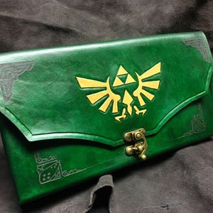 May include: A green leather journal with a gold embossed Triforce symbol. The journal has a flap closure with a metal clasp.