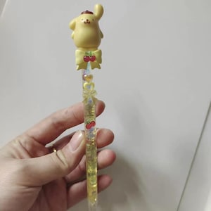 May include: A yellow pen with a cartoon dog character and a red hat. The pen has a yellow bow with red cherries and a clear barrel filled with yellow liquid and glitter. The pen is held by a person.