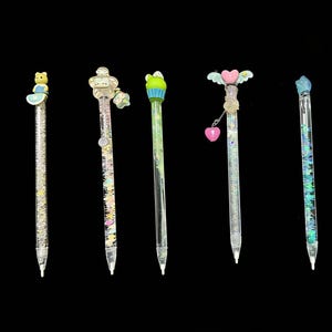 May include: Six decorative pens with clear barrels filled with glitter and small objects. Each pen has a different topper: a bear, a robot, a frog, a winged heart, a blue gem, and a cupcake. The pens are on a black background.