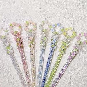 Puede incluir: Un conjunto de siete palillos decorativos para el cabello. Cada palillo presenta un diseño de corona floral en varios colores pastel, incluyendo rosa, verde y azul. Los tallos son transparentes con detalles de purpurina y un lazo decorativo.