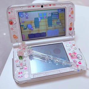 May include: White Nintendo 3DS handheld game console with a clear protective case decorated with pink and white stickers. A clear stylus with pink floral accents rests on the bottom screen. The top screen shows a colourful game.