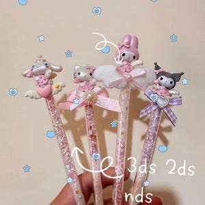 May include: Four decorative pens with cartoon character toppers. Each pen has a clear barrel filled with glitter. The characters include a white dog, Hello Kitty, a pink bunny, and a black cat. The pens are pink, white, and purple. The text "3ds 2ds nds" is visible.