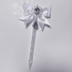 May include: A white pen with a decorative bow and jewel embellishments. The bow is made of satin-like material and features a large, ornate jewel in the centre. A teardrop-shaped jewel hangs below the centre jewel. The pen's barrel is filled with sparkling glitter.