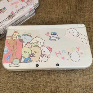 Puede incluir: Consola Nintendo 3DS XL blanca con un diseño colorido que presenta personajes de dibujos animados y la palabra "Happy". Los personajes están viajando e incluyen un oso, un pingüino y un gato.
