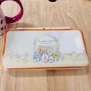 May include: A rectangular, off-white case with an orange border. The case features a floral design and the text "Sumikkogurashi". The case has a cartoon illustration of several characters.
