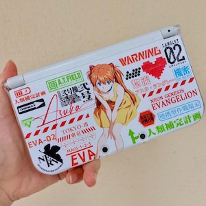 May include: White handheld gaming console with a vibrant anime design. The console features a character with orange hair and a yellow dress. Text includes "EVA-02", "Neon Genesis Evangelion", and Japanese characters.