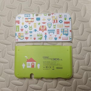 May include: A New Nintendo 3DS XL console with a white top cover featuring a colorful pattern of various objects. The bottom cover is lime green with a house graphic and the text "new: Nintendo 3DS XL".