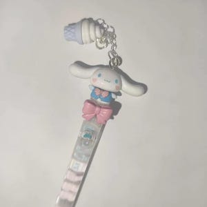 May include: A clear pen with a cartoon character design. The pen features a white and blue character with a pink bow, a blue bow tie, and a small ice cream cone charm. The pen's barrel has a pastel colour scheme.