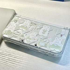 May include: A white Nintendo Switch console with a clear protective case decorated with various pastel-colored stickers featuring cartoon animals and other designs.