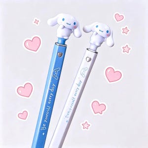 May include: Two pens, one blue and one white, each topped with a white cartoon character. The blue pen has the text "Be yourself every day" and a cartoon character. The white pen has the text "Treat yourself every day" and a cartoon character. Pink heart and star stickers are scattered around the pens.