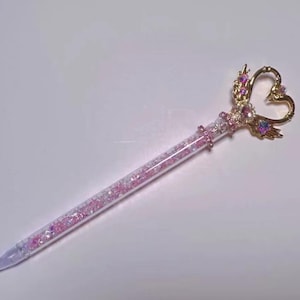 May include: A decorative pen with a clear barrel filled with pink glitter. The pen has a gold-coloured heart-shaped top adorned with pink and purple gems. The pen is designed with a pointed tip.