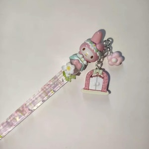 May include: A decorative pen featuring a pink My Melody character, a pink flower, and a pink door charm. The pen's shaft is clear with a pink and purple pattern. The pen is adorned with white flowers.