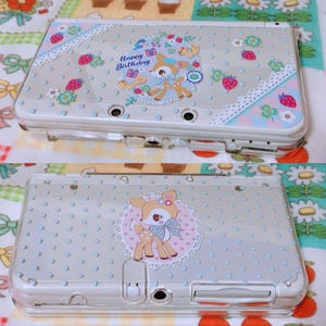 May include: A clear plastic case for a Nintendo 3DS with a light blue and white polka dot pattern. The case has a design of a cartoon deer with a bow on its head and the words "Happy Birthday" in a circle around it.