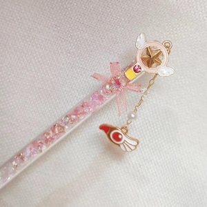May include: A pink and gold wand with a star-shaped charm, wings, and a dangling charm. The wand is filled with pink glitter and has a pink bow. The charm is red, white, and gold.