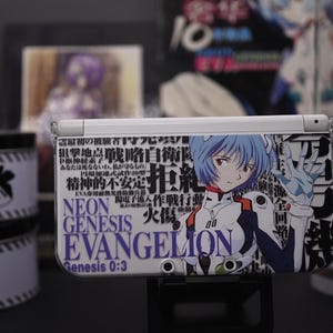May include: A white Nintendo 3DS console with a custom cover featuring an anime character with blue hair and the text "NEON GENESIS EVANGELION". The console is on a black stand, with other related merchandise in the background.