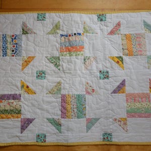 May include: A rectangular quilt featuring a white background with six central blocks composed of multi-colored fabric strips. Each block is surrounded by small triangular fabric pieces in various patterns and colors, finished with a yellow border.