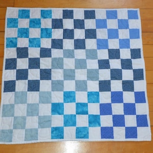 May include: A square quilt featuring a patchwork design of nine blocks, each composed of a four-by-four grid of alternating white and various blue fabric squares. The quilt has a white border and visible wavy machine quilting stitches throughout.