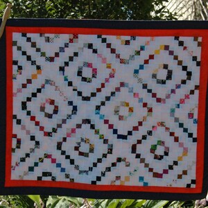 May include: A small, square quilt with a white background and a repeating diamond pattern. The diamonds are formed by small, colorful squares. The quilt has a red border and a dark blue outer border. It is hanging outside on a clothesline.
