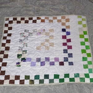 May include: A handmade quilt with a white center and a checkerboard border. The border features squares of brown, tan, green, and white. The quilt has a patchwork design with squares of various colors and patterns.