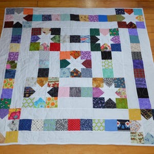 May include: A square patchwork quilt featuring a central star motif surrounded by smaller star blocks and a border of multi-colored fabric squares. The design incorporates a variety of patterned and solid-colored textiles separated by white fabric strips.