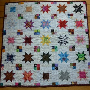 May include: A square patchwork quilt featuring a white background with a repeating pattern of multi-colored eight-pointed stars and small four-patch squares. The quilt is finished with a dark maroon border and displays intricate wavy quilting stitches.