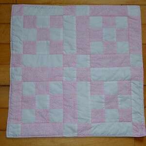 May include: A square quilted blanket featuring a repeating patchwork pattern of solid white fabric squares and pink fabric squares with a fine white floral print. The quilt is constructed with visible stitching lines and a finished white border edge.