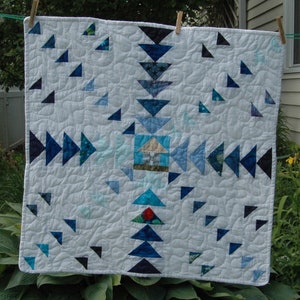 May include: A square, quilted blanket with a light blue background and a pattern of blue and teal triangles. A small house design is in the center. The quilt is hanging on a clothesline with wooden clothespins.
