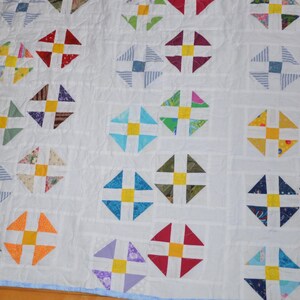 May include: A white quilt featuring a repeating pattern of multi-colored fabric squares arranged in a pinwheel design. Each pinwheel block contains four triangular segments surrounding a solid yellow center square, all stitched onto a white background.