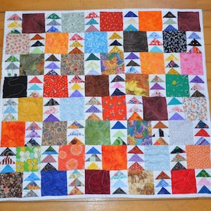 May include: A square patchwork quilt featuring a grid of multi-colored fabric squares and small triangular patterns. The quilt displays a variety of prints including floral, geometric, and solid colors in shades of red, blue, green, and orange.