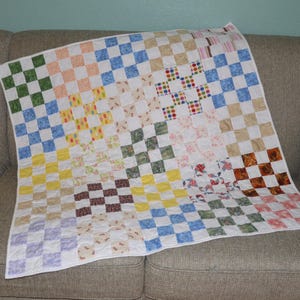 May include: A patchwork quilt featuring a grid of multi-colored fabric squares in various patterns including green, blue, yellow, brown, and pink. The quilt is constructed with white fabric sashing between the blocks and has a white border.