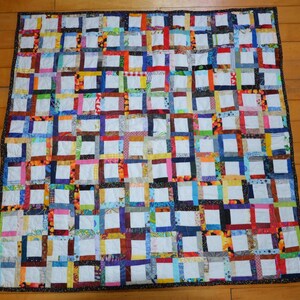 May include: A square patchwork quilt featuring a grid of white fabric squares surrounded by a variety of colorful, patterned fabric strips in shades of red, blue, yellow, green, and orange, finished with a dark, speckled fabric border.