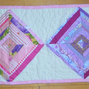 May include: A rectangular quilted table runner featuring two large diamond-shaped patchwork blocks centered on a cream-colored background. The left diamond uses pink and floral fabrics, while the right diamond uses purple and patterned fabrics.