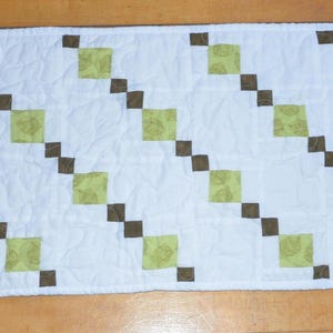 May include: A rectangular quilted table runner featuring a white background with a repeating diagonal pattern of small dark brown squares and larger light green squares. The fabric has a visible stippled quilting texture across the entire surface.