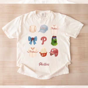 May include: Cream-colored t-shirt featuring a watercolor design of baseball-related icons, including a baseball, cap, jersey, bow, the letter "P", a mascot, a sundae, and a helmet. The word "Phillies" is printed below the icons. The text "Design #1" is at the bottom.