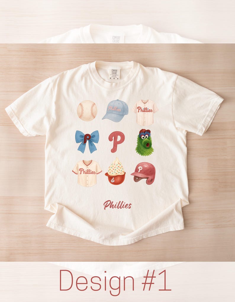 May include: Cream-colored t-shirt featuring a watercolor design of baseball-related icons, including a baseball, cap, jersey, bow, the letter "P", a mascot, a sundae, and a helmet. The word "Phillies" is printed below the icons. The text "Design #1" is at the bottom.