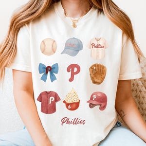 May include: Cream-colored t-shirt featuring baseball-themed illustrations. The design includes a baseball, a baseball cap, a jersey, a bow, the letter "P", a baseball glove, a shirt, a sundae, and a helmet. The word "Phillies" is written at the bottom.